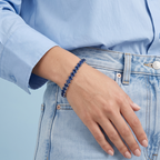 A close-up of a person wearing a Selforia India dark blue beaded bracelet on their wrist, styled with a light blue button-down shirt and light-wash denim jeans against a solid light blue background.