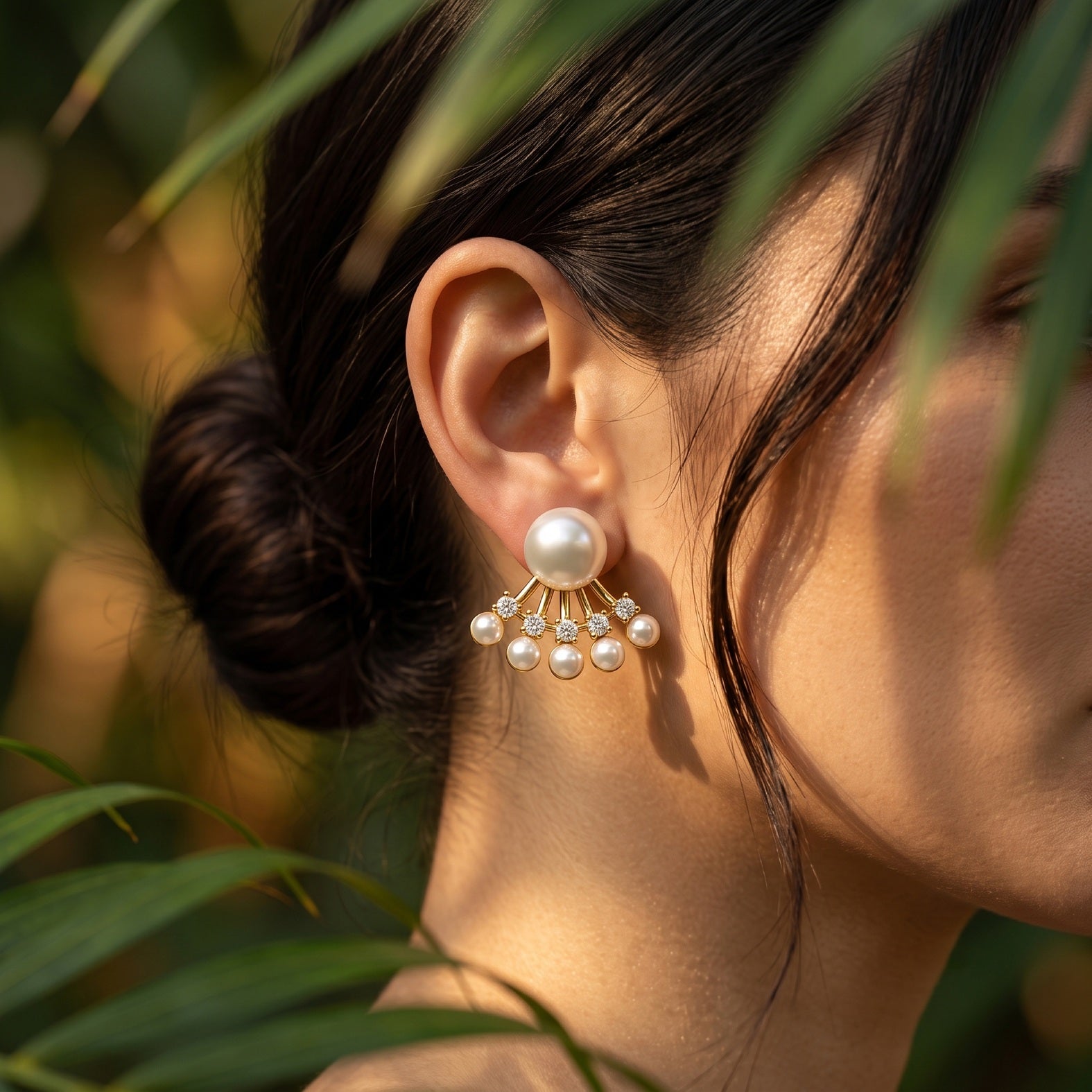 A close-up of a woman's ear wearing a dangling pearl earring with a large round pearl and smaller pearl clusters on a gold-tone setting, photographed outdoors with blurred green leaves in the background.