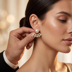 A close-up lifestyle shot of a woman wearing a gold and pearl fan-shaped stud earring, with a hand adjusting it on her ear. The background is a soft, warm, blurred bokeh.