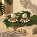 A pair of gold statement earrings with large white pearls and small clear stones, styled on green tropical leaves over a beige stone surface in warm natural light.