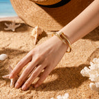 A Selforia Gold Nail bangle bracelet with a textured, hammered finish and a small row of clear stones, worn on a woman's wrist in a sandy beach setting with a straw hat and seashells.