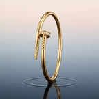 A Selforia Gold Nail hinged bangle bracelet styled like a nail, featuring small pavé-set clear stones, standing upright on a reflective dark surface against a soft gradient background.