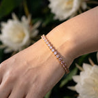 A close-up of a person's wrist wearing a delicate rose gold tennis bracelet with small clear stones, set against a soft-focus background of white flowers and green leaves.