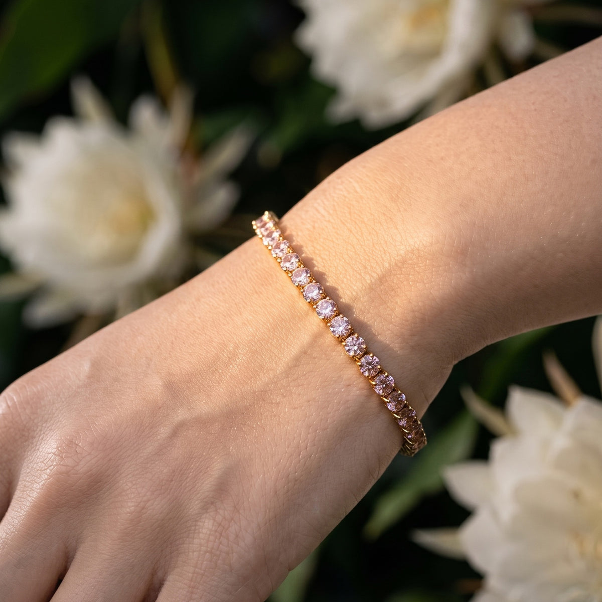 A close-up of a person's wrist wearing a delicate rose gold tennis bracelet with small clear stones, set against a soft-focus background of white flowers and green leaves.