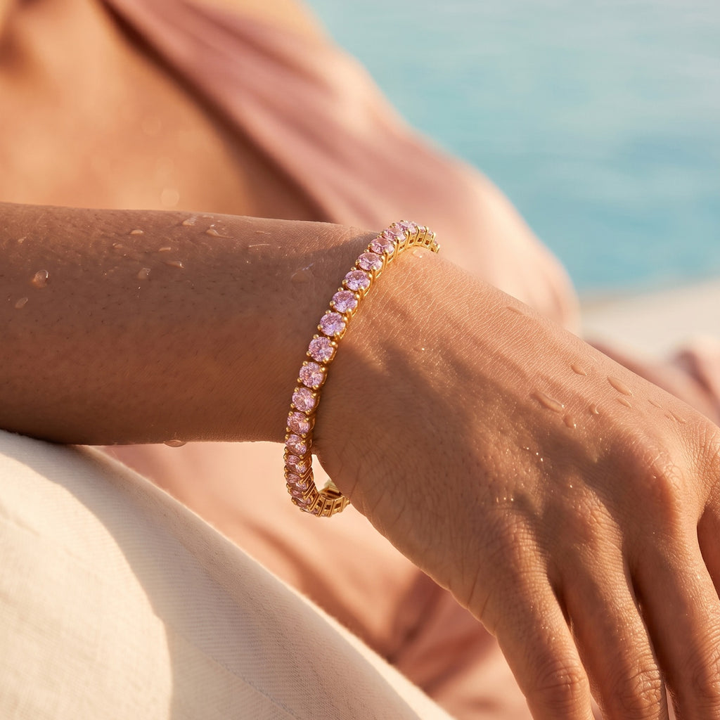 A close-up of a person's wrist wearing a Selforia India gold tone plated tennis bracelet with sparkling pink gemstones, set against a soft-focus background of a pink garment and blue water.