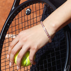 A close-up lifestyle shot of a woman's hand holding a tennis ball on a clay court while wearing a Selforia India gold toned plated tennis bracelet with pink gemstones.