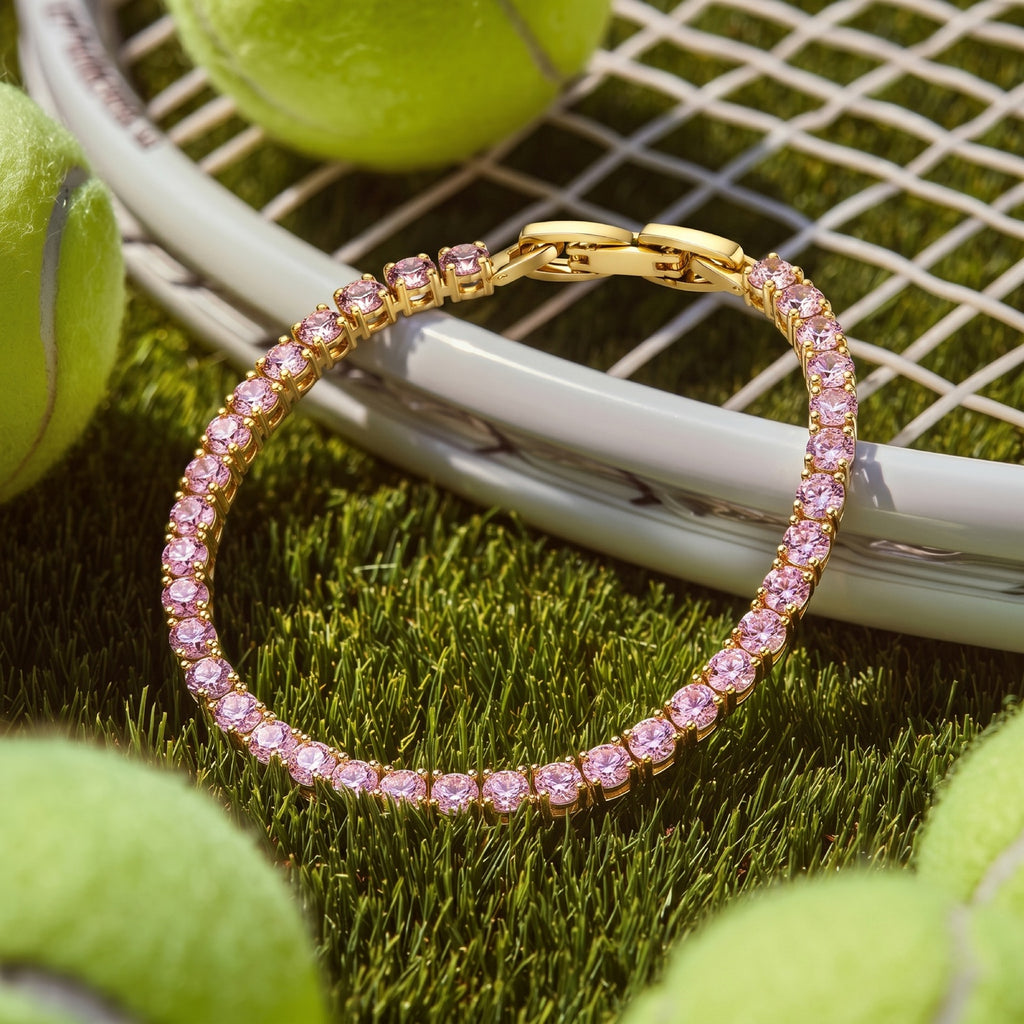 A Selforia gold toned plated tennis bracelet with pink gemstones, styled on green grass with tennis rackets and tennis balls.