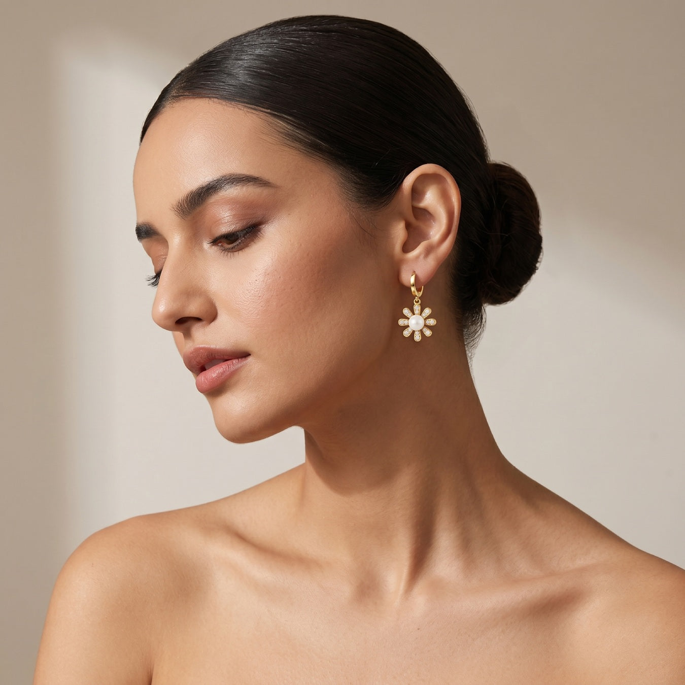 A woman with her hair in a low bun models a delicate gold dangle earring featuring a white flower with small clear stones, set against a warm beige background.