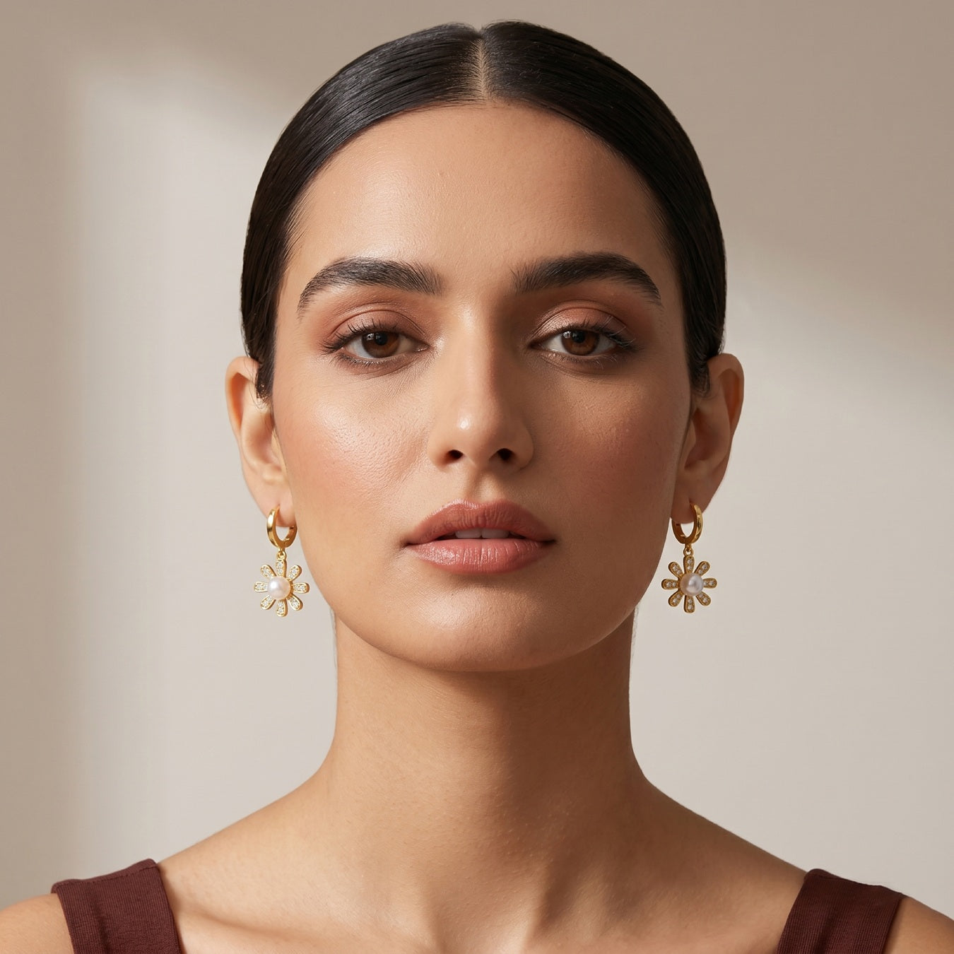 A close-up portrait of a woman wearing gold dangle earrings with a floral or sunburst design, set against a soft beige background.