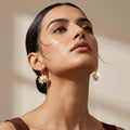 A close-up lifestyle shot of a woman wearing gold daisy-shaped drop earrings with white flower petals, set against a warm beige background with soft shadows.