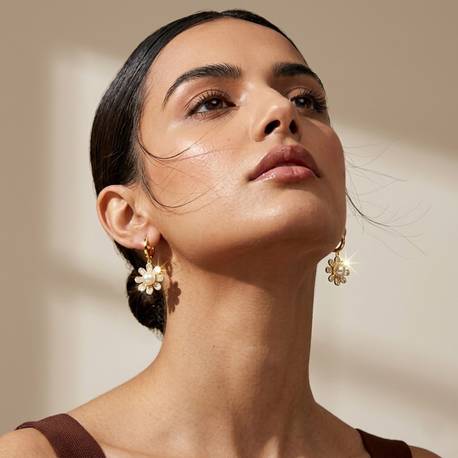 A close-up lifestyle shot of a woman wearing gold daisy-shaped drop earrings with white flower petals, set against a warm beige background with soft shadows.